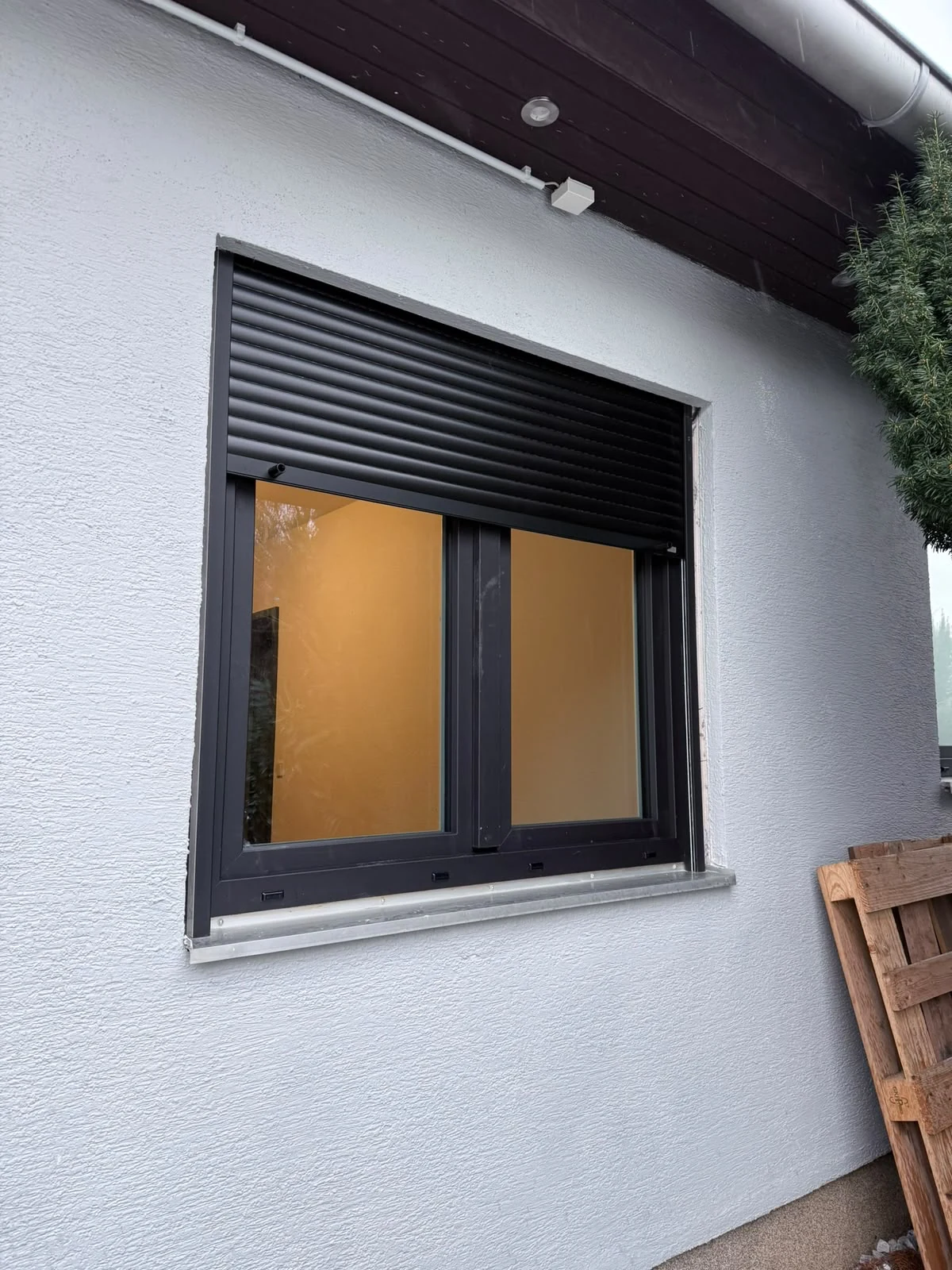 Fenster mit dunklem Rahmen und teilweise geschlossenem Rollladen an einer hellen Fassade.